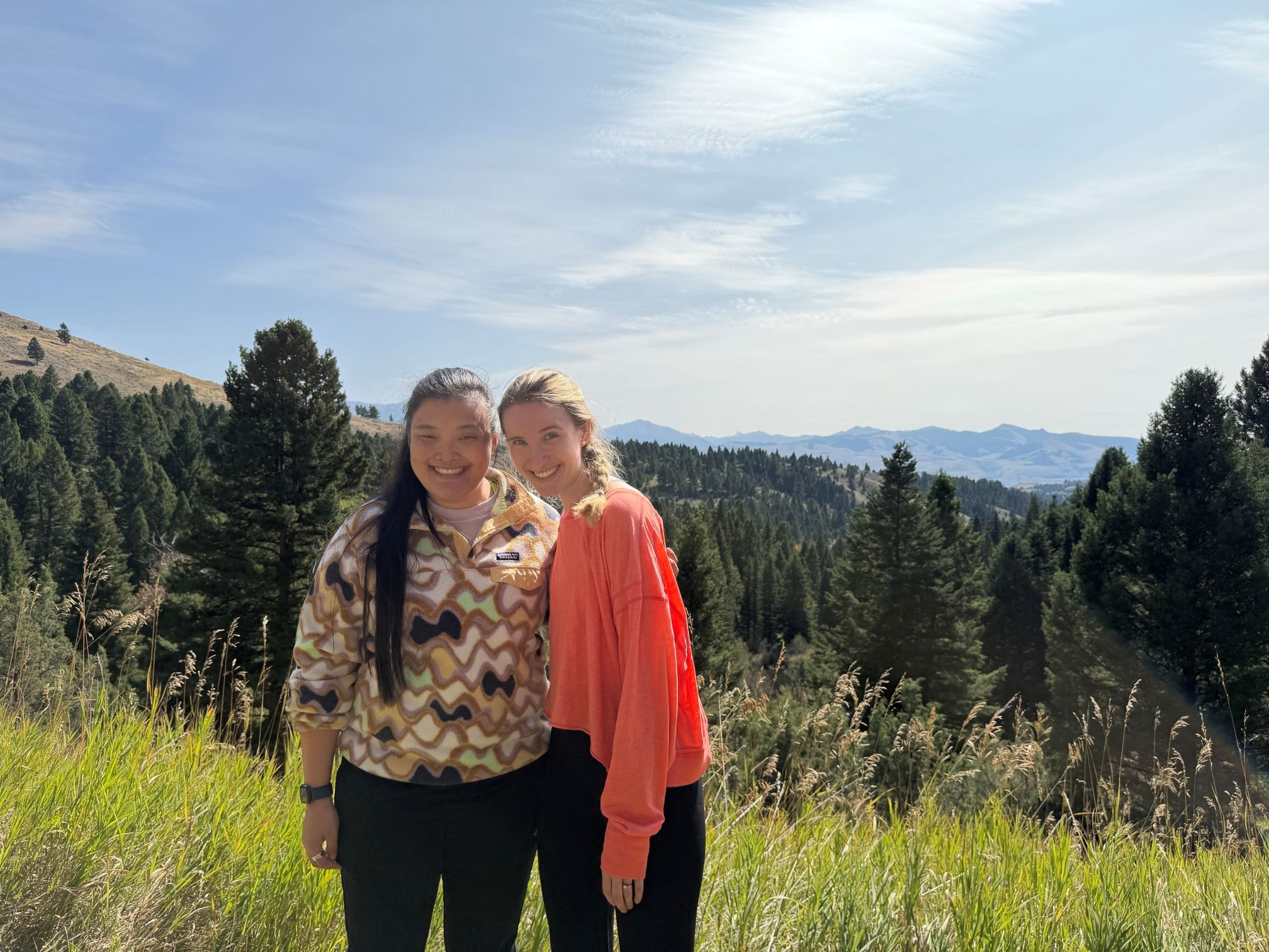 Wellness lead residents, Maddie du Pont, DO, class of 2027 & Holly Wilson, DO, class of 2028, stand together in athleisure wear on a hiking trip, with green grass, pine trees, and blue sky behind them.