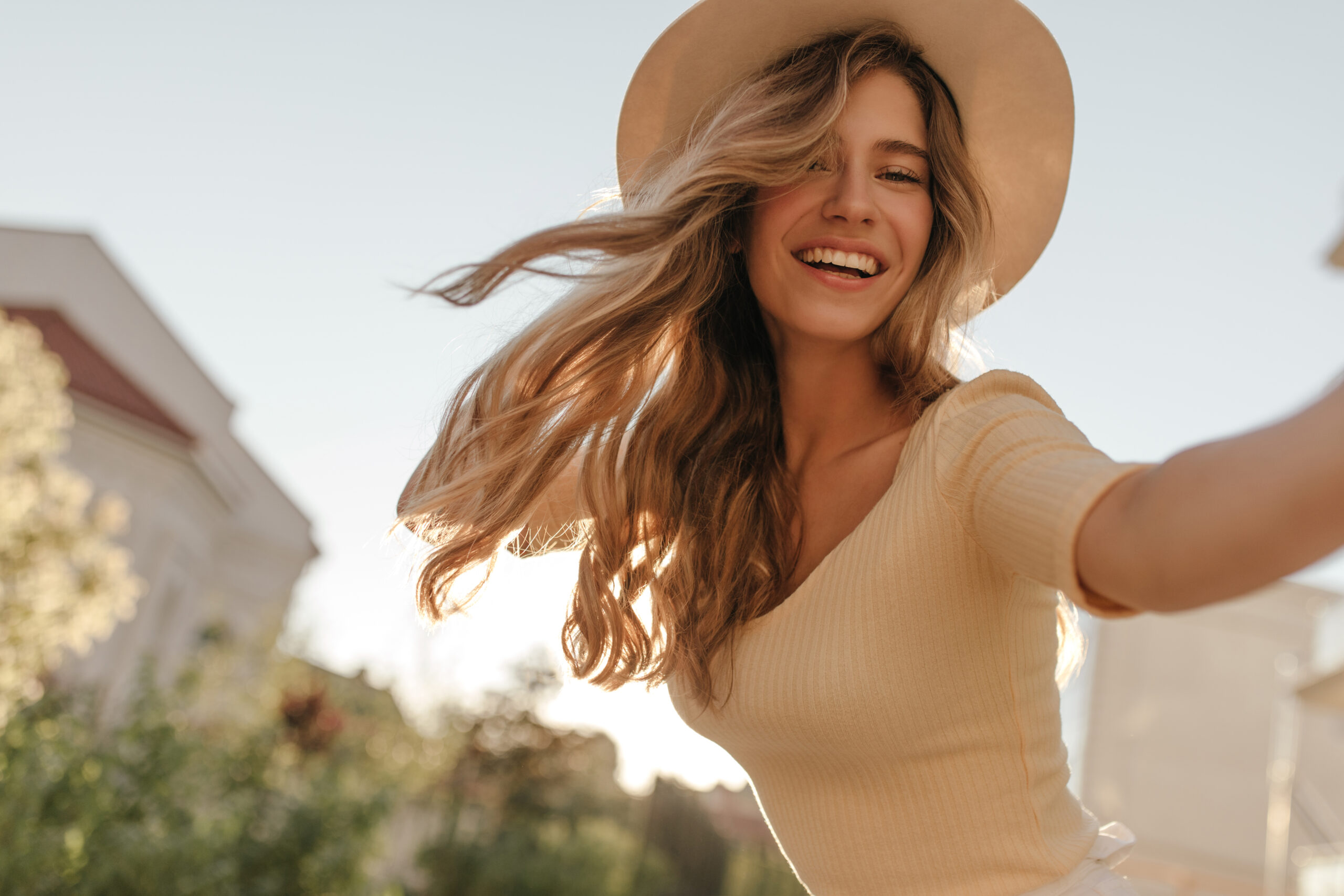 Happy young woman holding camera in hand close-up. Her hair is flying in wind, she is wearing hat. Outdoor photo.